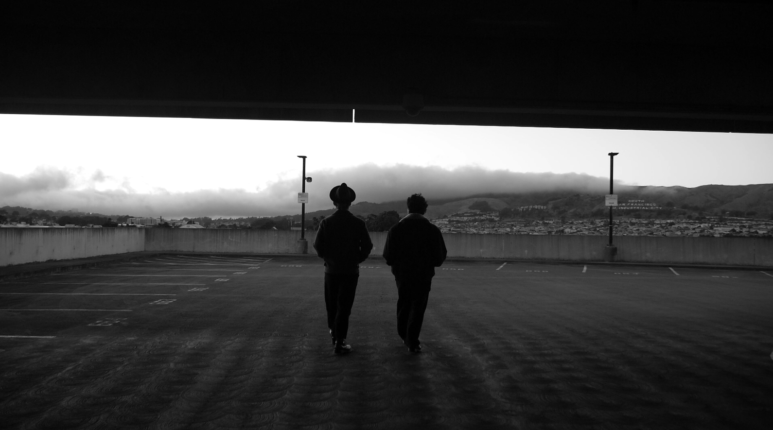 Two figures walk across a rooftop parking structure overlooking San Bruno